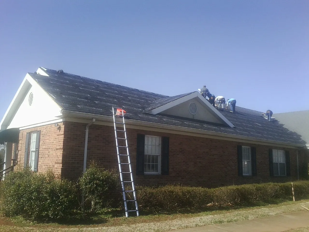 Crew tearing shingles off a brick ranch roof with a ladder set — Emergency Roof Repair work by our crew in Lynn Haven
